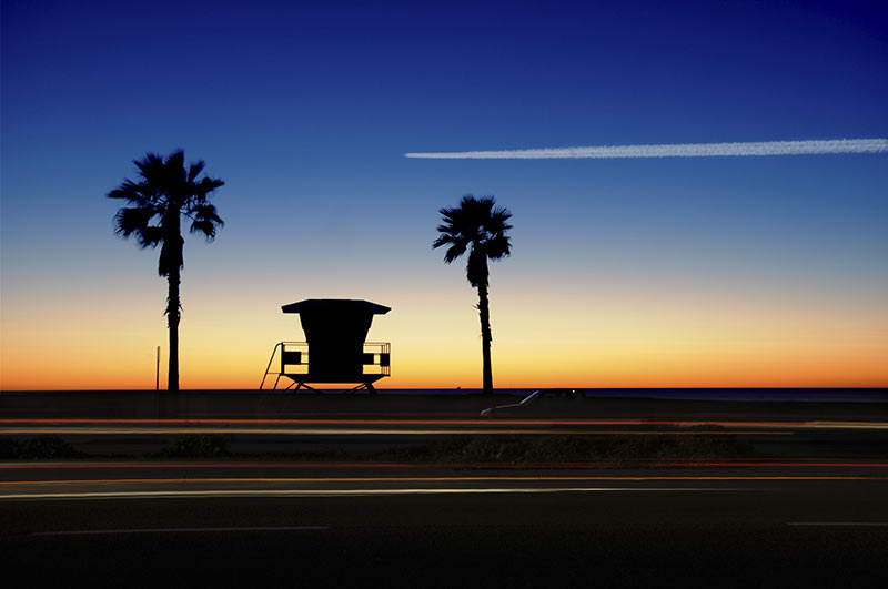 Image of a lifeguard tower and two palm trees silhouetted at sunset by the beach in Carlsbad, California as seen from the road.